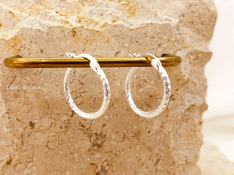 May include: Silver hoop earrings with a textured, twisted pattern. The earrings are suspended from a gold-coloured bar, set against a light-coloured stone backdrop. The brand name "TibiCollection" is visible.