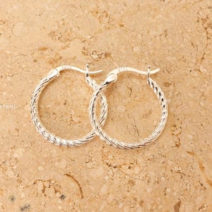 May include: A pair of silver hoop earrings with a textured, braided design. The earrings are displayed on a light brown, speckled surface. The earrings are a classic jewellery item.