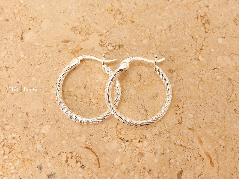 May include: A pair of silver hoop earrings with a textured, braided design. The earrings are displayed on a light brown, speckled surface. The earrings are a classic jewellery item.