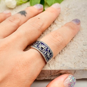 Può includere: Un anello d'argento con una fascia in smalto blu con un motivo floreale. L'anello è indossato su un dito.