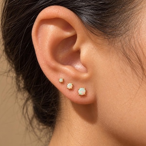 May include: Three round opal stud earrings set in gold-toned metal prongs, arranged in a line along the earlobe. Each earring features a smooth, iridescent white stone that displays subtle flashes of color, secured by a simple four-prong setting.