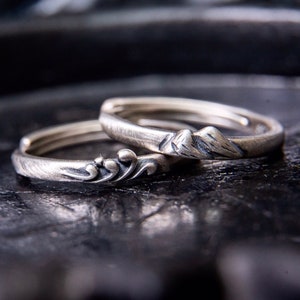 May include: Two silver rings with nature-inspired designs. One ring features a mountain range, while the other has an ornate, flowing pattern. The rings are set against a dark, textured background, highlighting their intricate details.