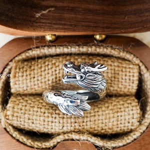 May include: A silver dragon ring with intricate details, displayed in a wooden box with a burlap lining. The ring features a dragon head and tail design. The ring is a unique piece of jewelry.