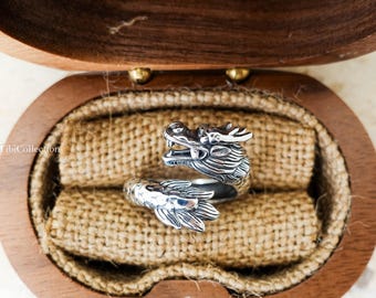 Può includere: Un anello a forma di drago in argento con dettagli intricati, esposto in una scatola di legno con fodera in juta. L'anello presenta un design a testa e coda di drago. Un gioiello unico.