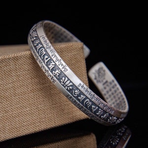 May include: Silver bracelet with a decorative design and engraved text in Tibetan script. The bracelet is open and adjustable.