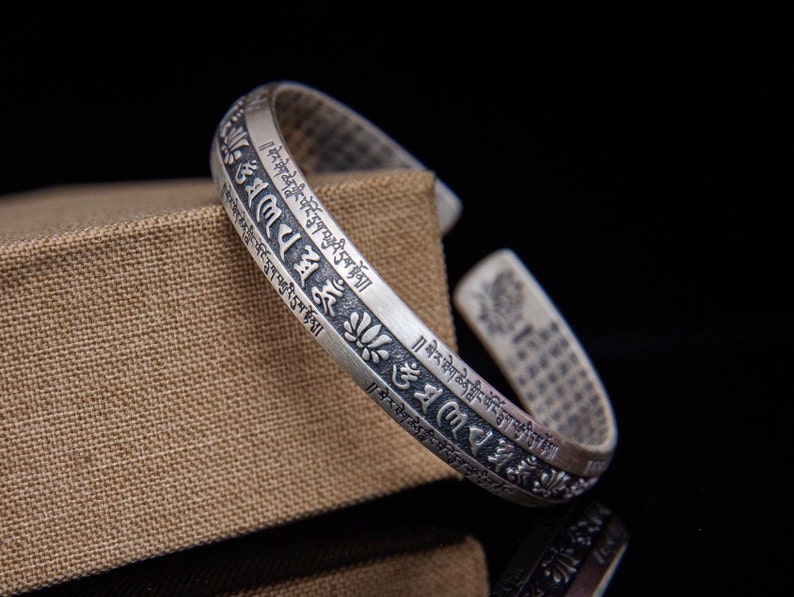 May include: Silver bracelet with a decorative design and engraved text in Tibetan script. The bracelet is open and adjustable.