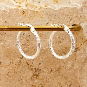 May include: Two silver hoop earrings with a textured, woven pattern. They are presented on a gold-coloured bar, set against a textured, beige backdrop. These earrings are a classic jewellery item.