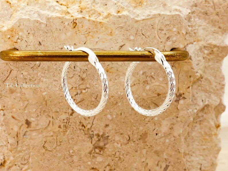 May include: Two silver hoop earrings with a textured, woven pattern. They are presented on a gold-coloured bar, set against a textured, beige backdrop. These earrings are a classic jewellery item.
