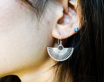 May include: Silver earrings with a fan-shaped design. The earrings are made of thin metal wires that are connected to form a circle in the centre. The earrings are hanging from a small hook.