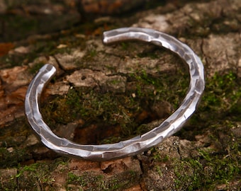 May include: A silver metal bracelet with a hammered texture. The bracelet is thick and has a rounded shape.