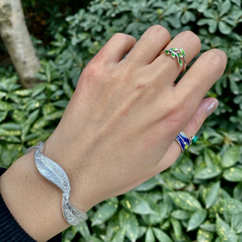 Pode incluir: Uma pulseira de prata com um design de folha e um anel de prata com folhas de esmalte verde. A joia est&aacute; na m&atilde;o de uma pessoa.