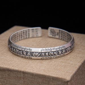 May include: Silver bracelet with a repeating inscription in Tibetan script. The bracelet is on a brown surface.