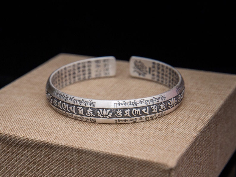 May include: Silver bracelet with a repeating inscription in Tibetan script. The bracelet is on a brown surface.