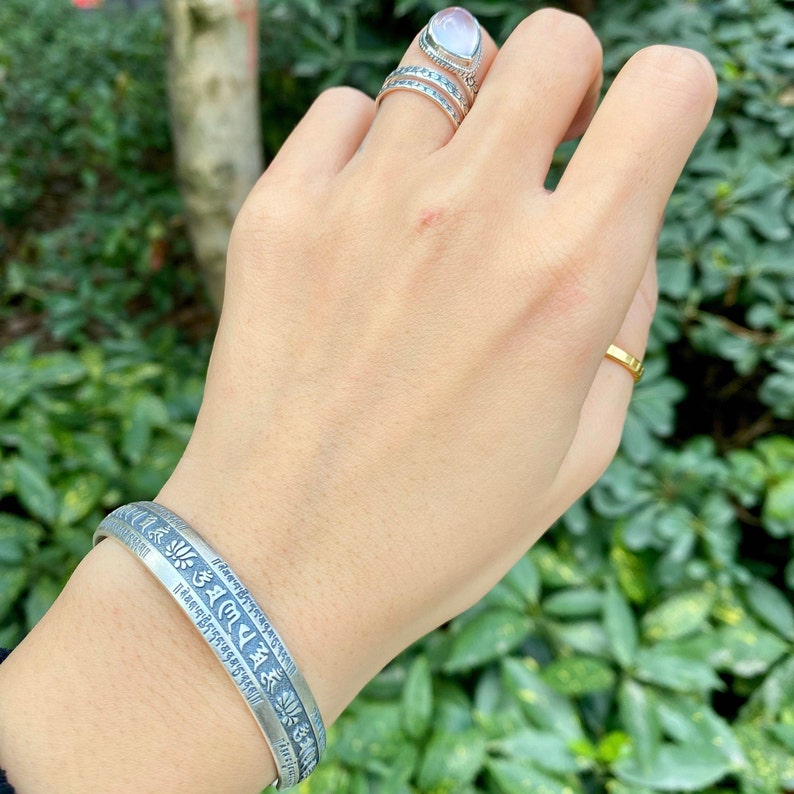 May include: A silver bracelet with a decorative inscription in Tibetan script. The bracelet is worn on a person's wrist.