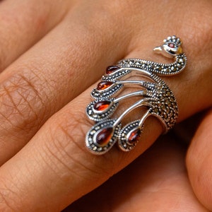 May include: A silver ring with a peacock design. The peacock's tail feathers are adorned with orange gemstones.