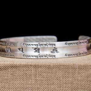 May include: A silver cuff bracelet with etched Tibetan script. The bracelet has a brushed metal finish and a curved design. The script is evenly spaced around the band. The bracelet is displayed on a neutral-toned surface.