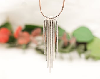 May include: A silver necklace with a long, thin, dangling chain made of multiple strands. The chain is silver and has a slightly textured surface. The necklace is photographed against a blurred background of green and pink.