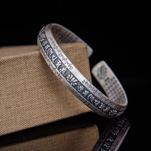 May include: Silver bracelet with an intricate design of Tibetan script and lotus flowers. The bracelet is open and adjustable.