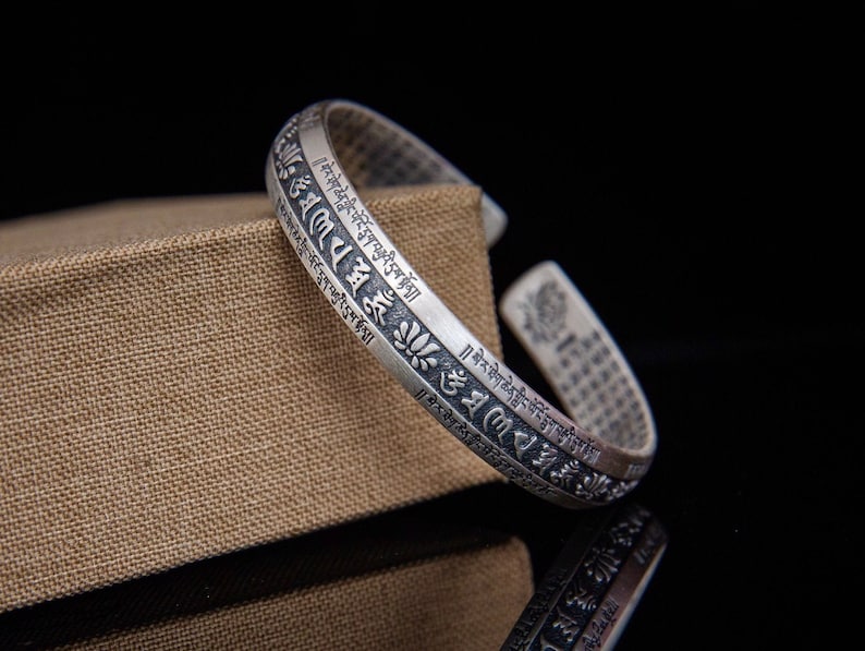 May include: Silver bracelet with an intricate design of Tibetan script and lotus flowers. The bracelet is open and adjustable.
