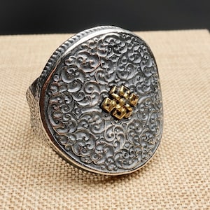 May include: A silver-toned ring with an ornate, swirling pattern and a gold-colored central design. The ring has a wide band and a detailed, textured surface. The gold design is a small, symmetrical shape.