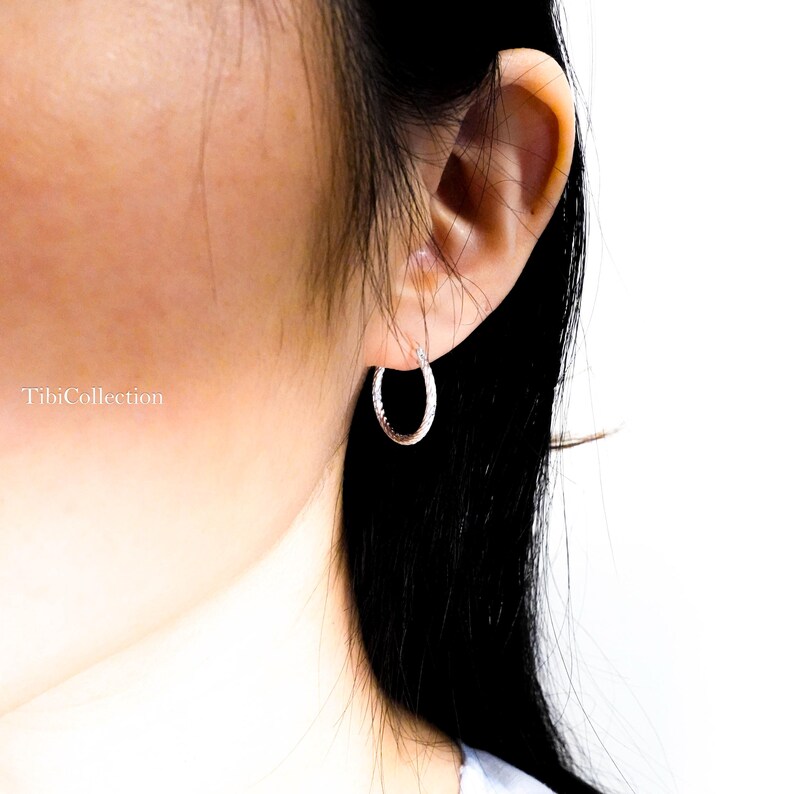 May include: A close-up of a silver hoop earring with a textured surface. The earring is worn on the ear of a person. The brand name "TibiCollection" is visible in the image.