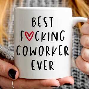 K&ouml;nnte beinhalten: Wei&szlig;e Keramik-Tasse mit dem Text "BEST FUCKING COWORKER EVER" in schwarzer Schrift. Ein rotes Herz ersetzt das "O" in "FUCKING". Die Tasse wird von einer Person gehalten.