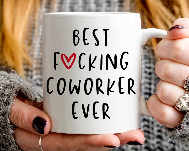 K&ouml;nnte beinhalten: Wei&szlig;e Keramik-Tasse mit dem Text "BEST FUCKING COWORKER EVER" in schwarzer Schrift. Ein rotes Herz ersetzt das "O" in "FUCKING". Die Tasse wird von einer Person gehalten.