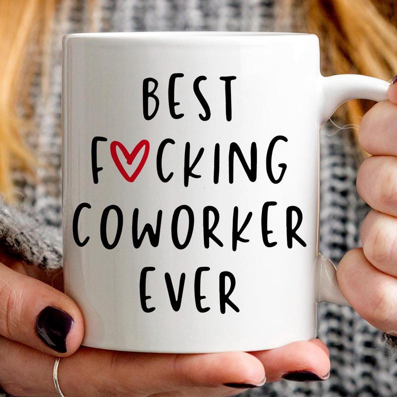 K&ouml;nnte beinhalten: Wei&szlig;e Keramiktasse mit dem Text "BEST F*CKING COWORKER EVER" in Schwarz, mit einem roten Herz, das das "O" in "F*CKING" ersetzt. Die Tasse wird von einer Person mit dunklem Nagellack gehalten.