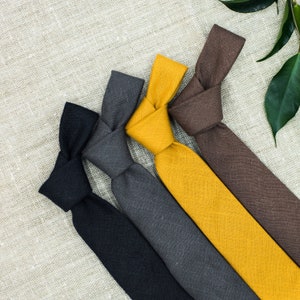 Puede incluir: Cinco corbatas de diferentes colores, incluyendo negro, gris, marrón y amarillo, dispuestas en forma de abanico sobre un fondo de lino beige.