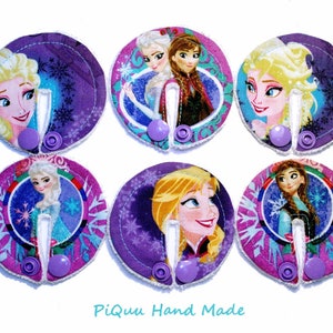 May include: Six purple and pink fabric circles with a white button in the center. Each circle features a different image of a Disney Frozen character, including Elsa and Anna. The text "Piquu Hand Made" is printed at the bottom of the image.