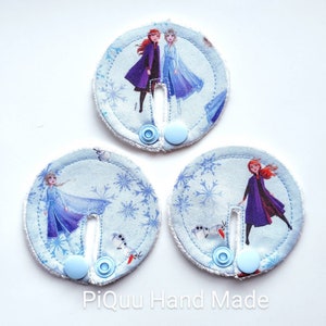 May include: Three round fabric appliques with a blue snowflake pattern and images of the characters Elsa and Anna from the movie Frozen. Each applique has a blue button and a slit in the center.
