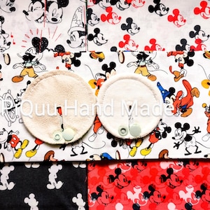 May include: Two round, beige nursing pads with snap closures are displayed on a fabric featuring a Mickey Mouse print. The fabric has a white background with Mickey Mouse in various poses and colours, along with black and red Mickey Mouse patterns.