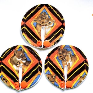 Könnte beinhalten: Drei runde Stoff-Stillkissen mit einem bunten geometrischen Muster und einem Bild von zwei Löwenjungen. Die Kissen haben eine weiße Mitte mit einem Druckknopfverschluss.