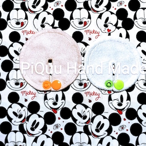 May include: Two circular absorbent pads, one pink and one white, each with a slit and button detail, are shown against a white fabric patterned with black and white Mickey Mouse faces. The text "PiQu Hand Made" is visible.