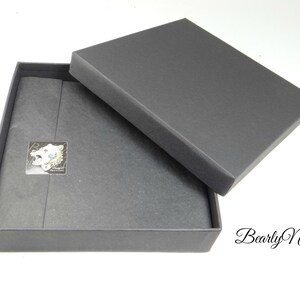 May include: A black square box with a lid. The box is lined with black tissue paper. A small white sticker with a black and white image of a bear's head is on the tissue paper. The sticker says "Bearly N" in white text.