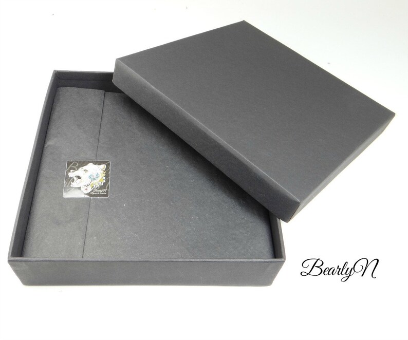 May include: A black square box with a lid. The box is lined with black tissue paper. A small white sticker with a black and white image of a bear's head is on the tissue paper. The sticker says "Bearly N" in white text.