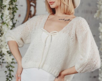 Può includere: Una donna indossa un maglione di lana bianco con un dettaglio a fiocco, una gonna bianca e un cappello bianco. È in piedi davanti a un muro di mattoni bianchi con fiori bianchi appesi al soffitto.