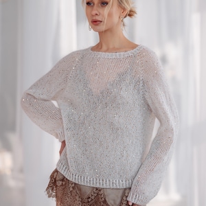 Könnte beinhalten: Ein hellfarbener Langarmpullover mit rundem Ausschnitt und funkelnden Verzierungen. Der Pullover wird mit einem braunen Spitzenrock und einer Paillettenhose kombiniert. Das Outfit ist für besondere Anlässe geeignet.