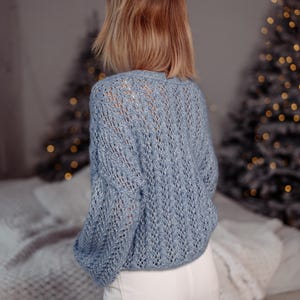 May include: A light blue knitted sweater with a delicate openwork pattern. The sweater has a relaxed fit and long sleeves.