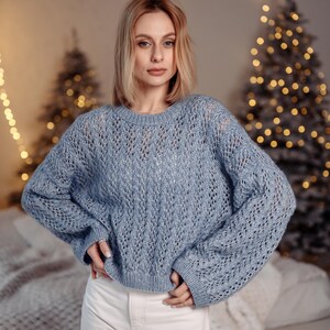 May include: A woman wearing a light blue knitted sweater with a delicate openwork pattern. She is standing in front of a Christmas tree with white lights.