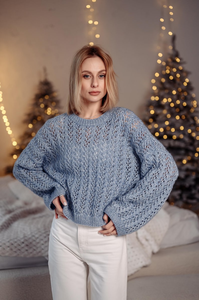 May include: A woman wearing a light blue knitted sweater with a delicate openwork pattern. She is standing in front of a Christmas tree with white lights.