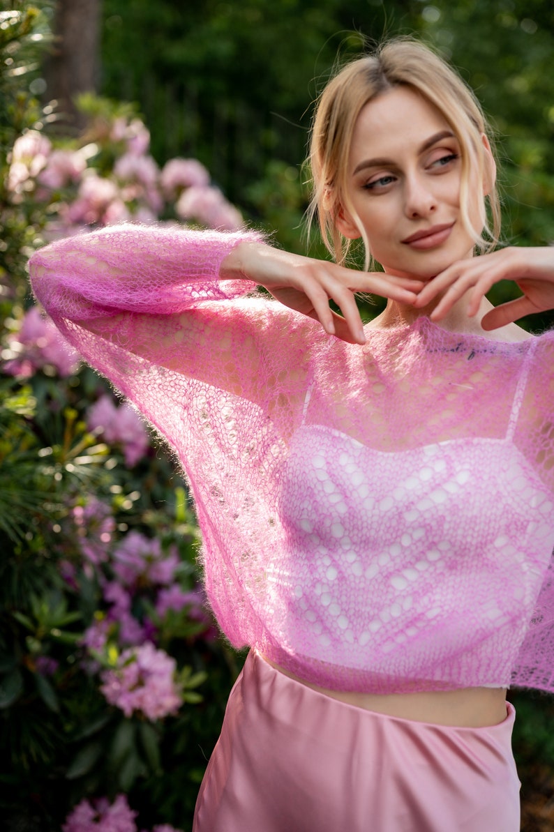 May include: A pink, lacy, lightweight sweater with a loose fit. The sweater is sheer and has a delicate, open-knit design.