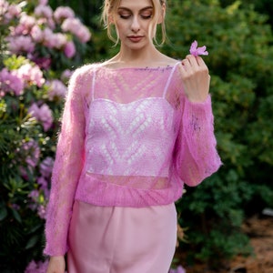 May include: A woman wearing a pink, lacy, knitted sweater over a white, lacy top and a pink skirt. She is holding a pink flower in her hand.