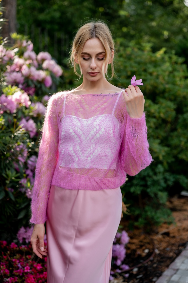 May include: A woman wearing a pink, lacy, knitted sweater over a white, lacy top and a pink skirt. She is holding a pink flower in her hand.