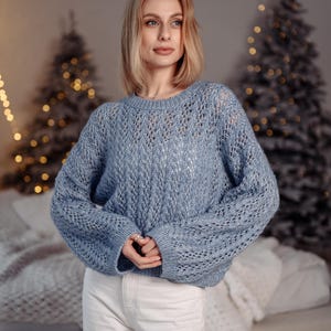 May include: A woman wearing a light blue knitted sweater with a delicate openwork pattern. She is standing in front of a Christmas tree with white lights.