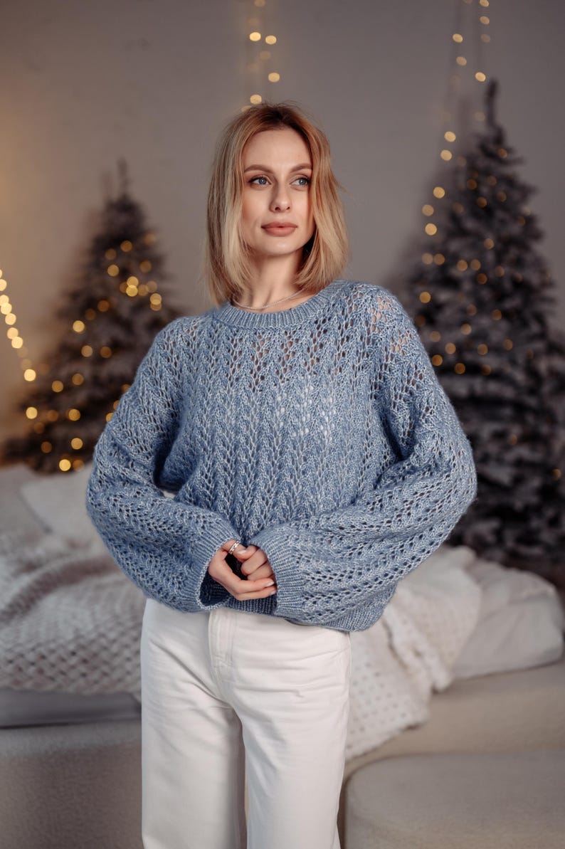 May include: A woman wearing a light blue knitted sweater with a delicate openwork pattern. She is standing in front of a Christmas tree with white lights.
