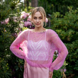 May include: A woman wearing a pink, sheer, knitted sweater over a white, lacy top and a pink skirt. She is standing in a garden with green foliage and pink flowers.
