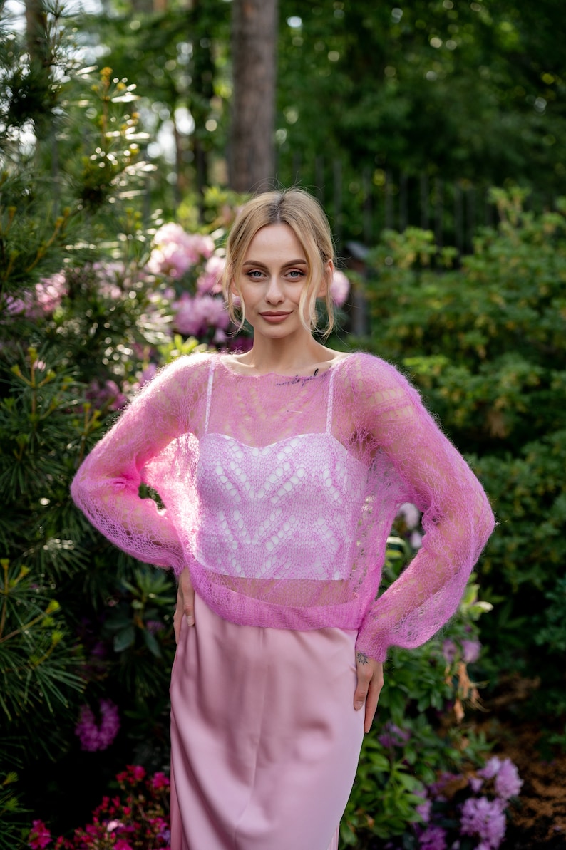 May include: A woman wearing a pink, sheer, knitted sweater over a white, lacy top and a pink skirt. She is standing in a garden with green foliage and pink flowers.