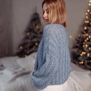 May include: A woman wearing a light blue knitted sweater with a delicate openwork pattern. The sweater has long sleeves and a relaxed fit. She is wearing white pants and is standing in front of a blurred background with Christmas lights.