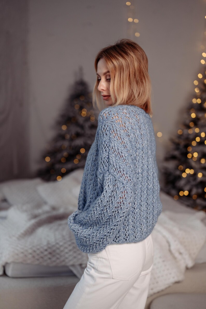 May include: A woman wearing a light blue knitted sweater with a delicate openwork pattern. The sweater has long sleeves and a relaxed fit. She is wearing white pants and is standing in front of a blurred background with Christmas lights.